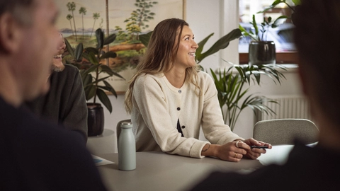 En gruppe mennesker deltager i en arbejdsbeskrivelse, hvor en smilende kvinde i en lys trøje sidder ved et bord med planter i baggrunden.